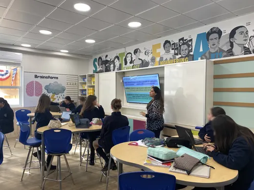 Cote presenta en un laboratorio mientras sus alumnas trabajan en mesas con computadores; las caras de las alumnas están difuminadas