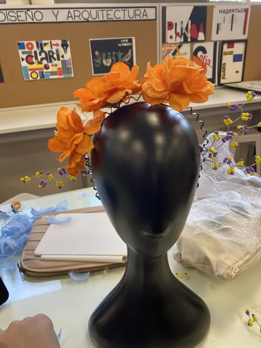 Tocado de flores naranjas sobre una cabeza de maniquí negra, frente a un panel de Diseño y Arquitectura